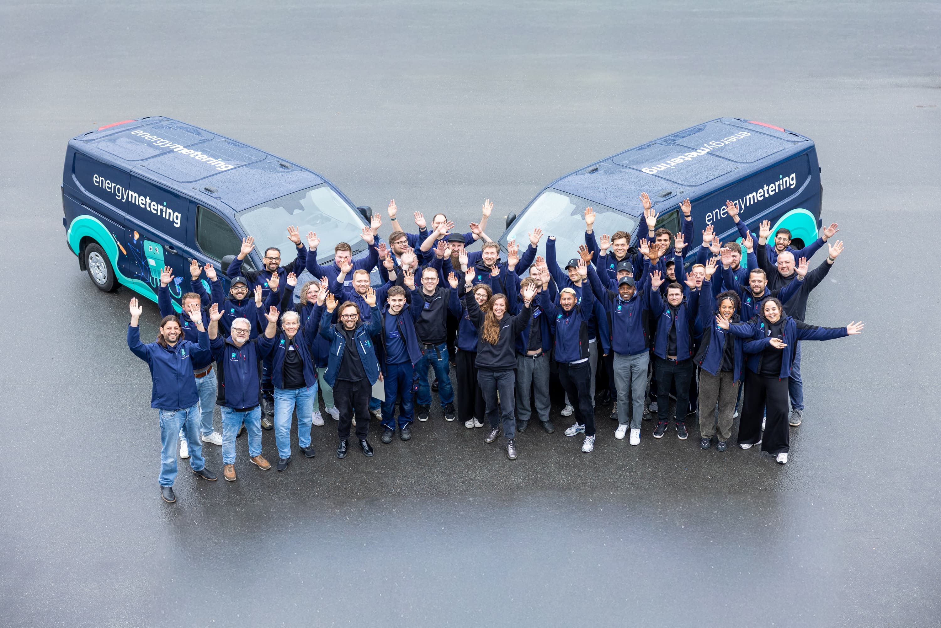 A group of people wearing matching jackets cheerfully pose in front of two branded vans in a heart formation, outdoors.