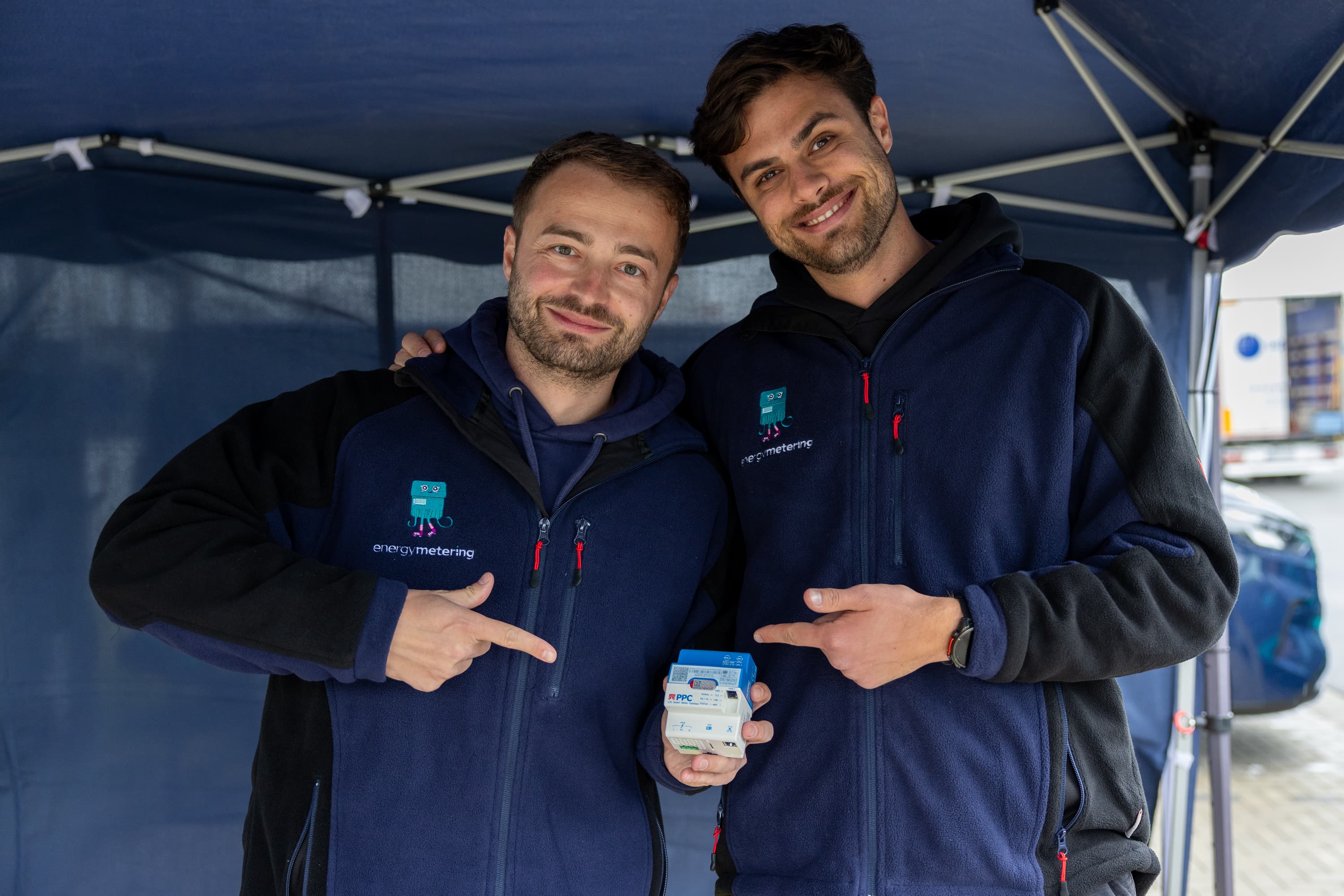Two men in matching blue jackets smiling and pointing at a smart meter gateway, standing under a canopy.